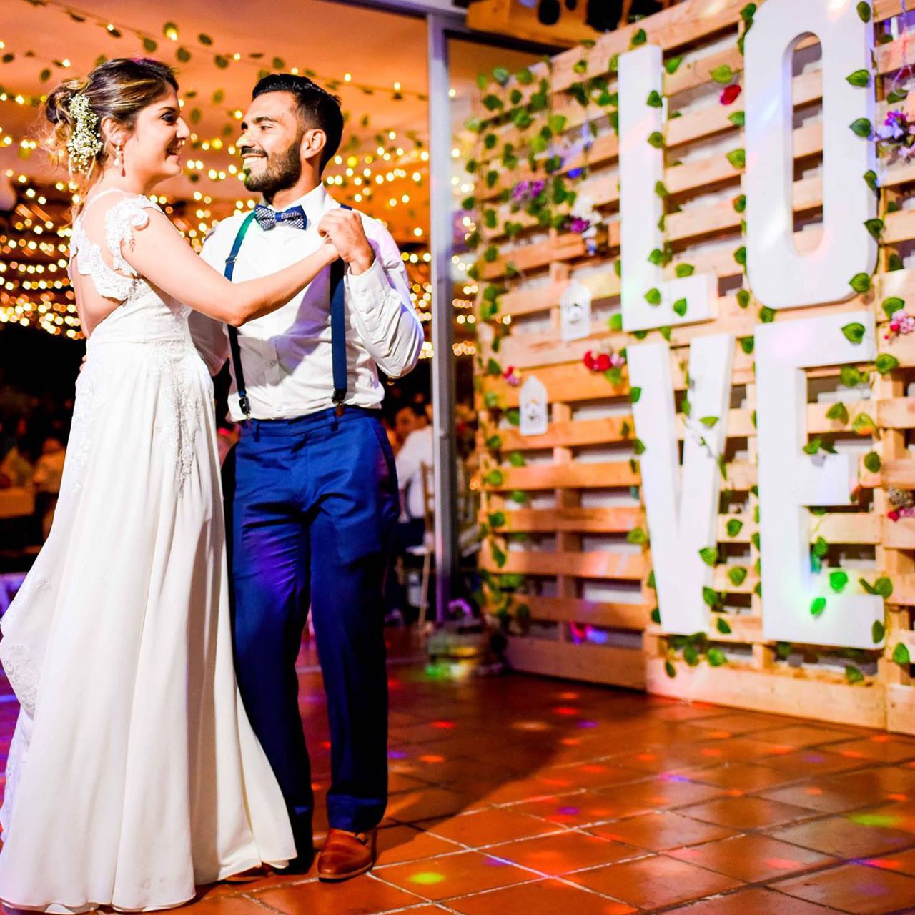 Pareja de novios bailando su primer vals frente a pared de pallets con letras LOVE y enredaderas