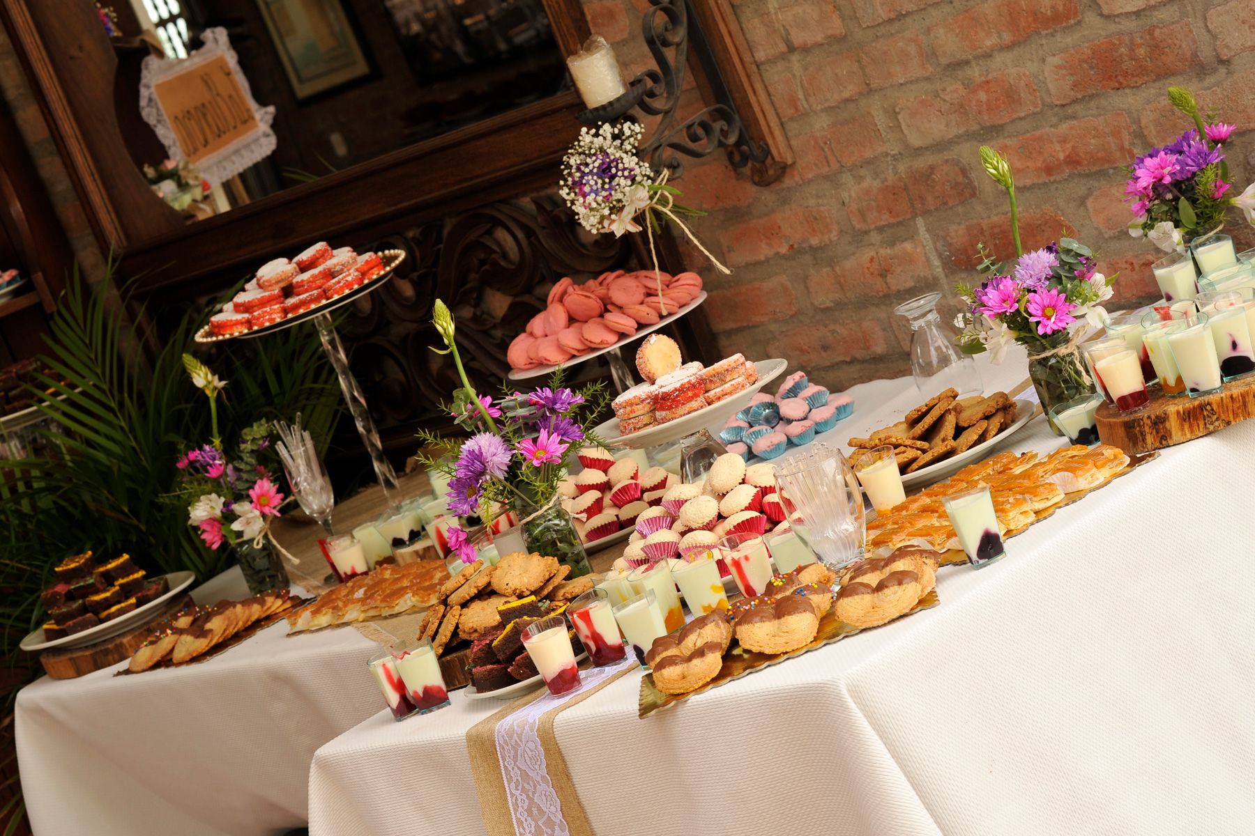 Mesa dulce con macarons, galletas y postres variados decorada con flores frescas junto a pared de ladrillo