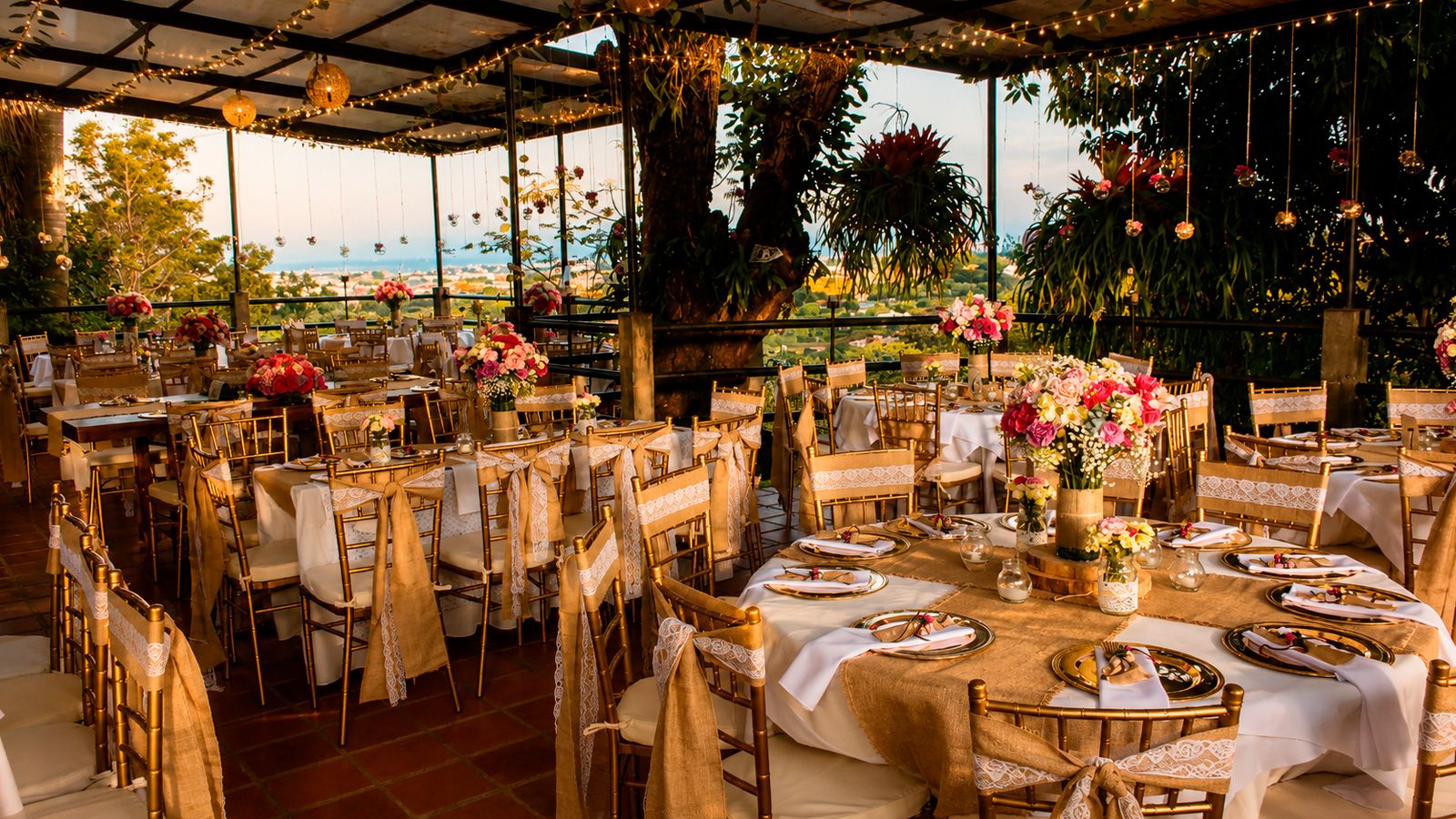 Salón de Casa La Eleonora con mesas redondas en sillas tiffany doradas, centros de flores fucsia y luces colgantes al atardecer