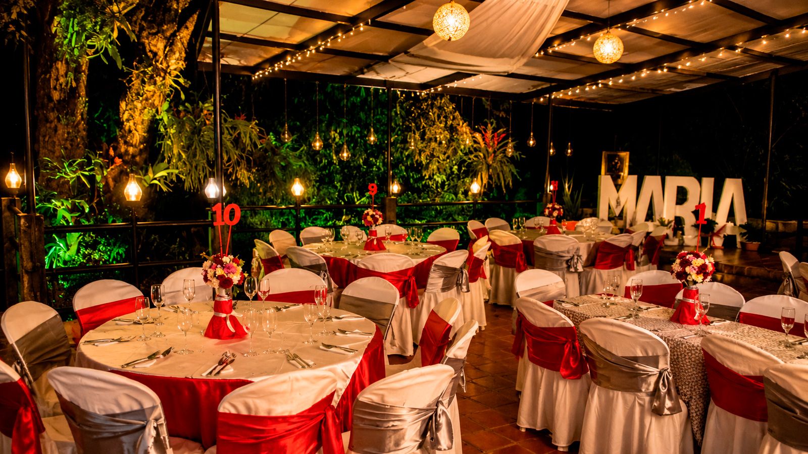Salón de Casa La Eleonora decorado en rojo y blanco para quinceañera, con letras iluminadas MARIA, lámparas Edison y vegetación