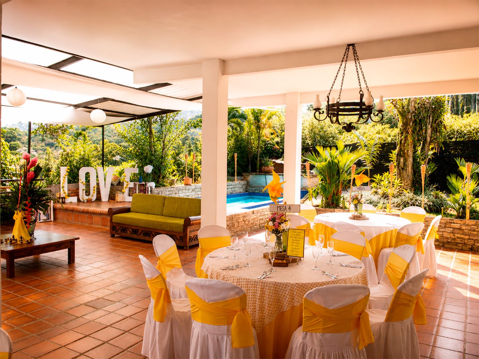 Terraza de Casa La Eleonora decorada con mesas en sillas blancas con lazos amarillos, letrero LOVE y piscina al fondo